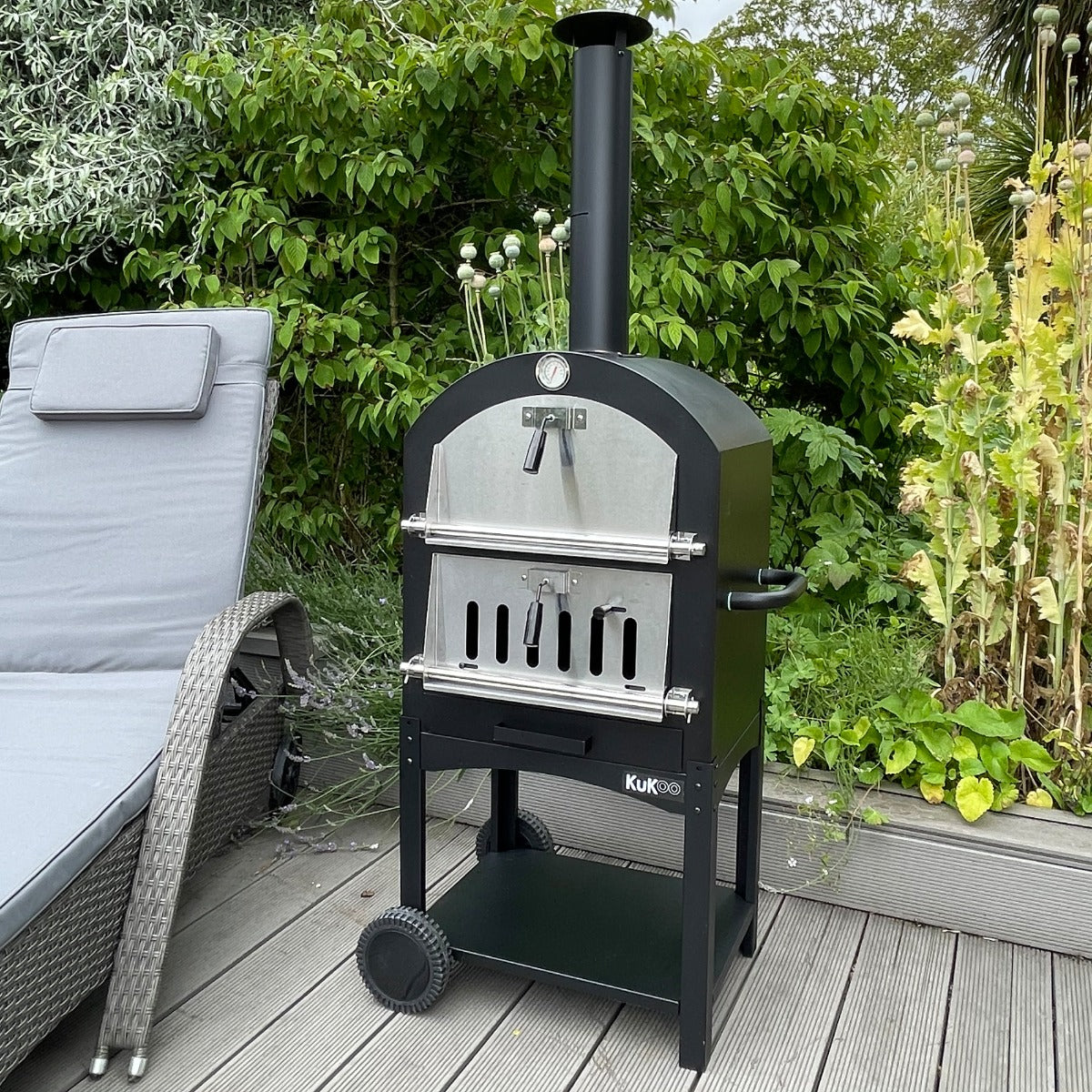 Black KuKoo outdoor pizza oven with stainless steel door, positioned on a wooden deck surrounded by greenery and a lounge chair
