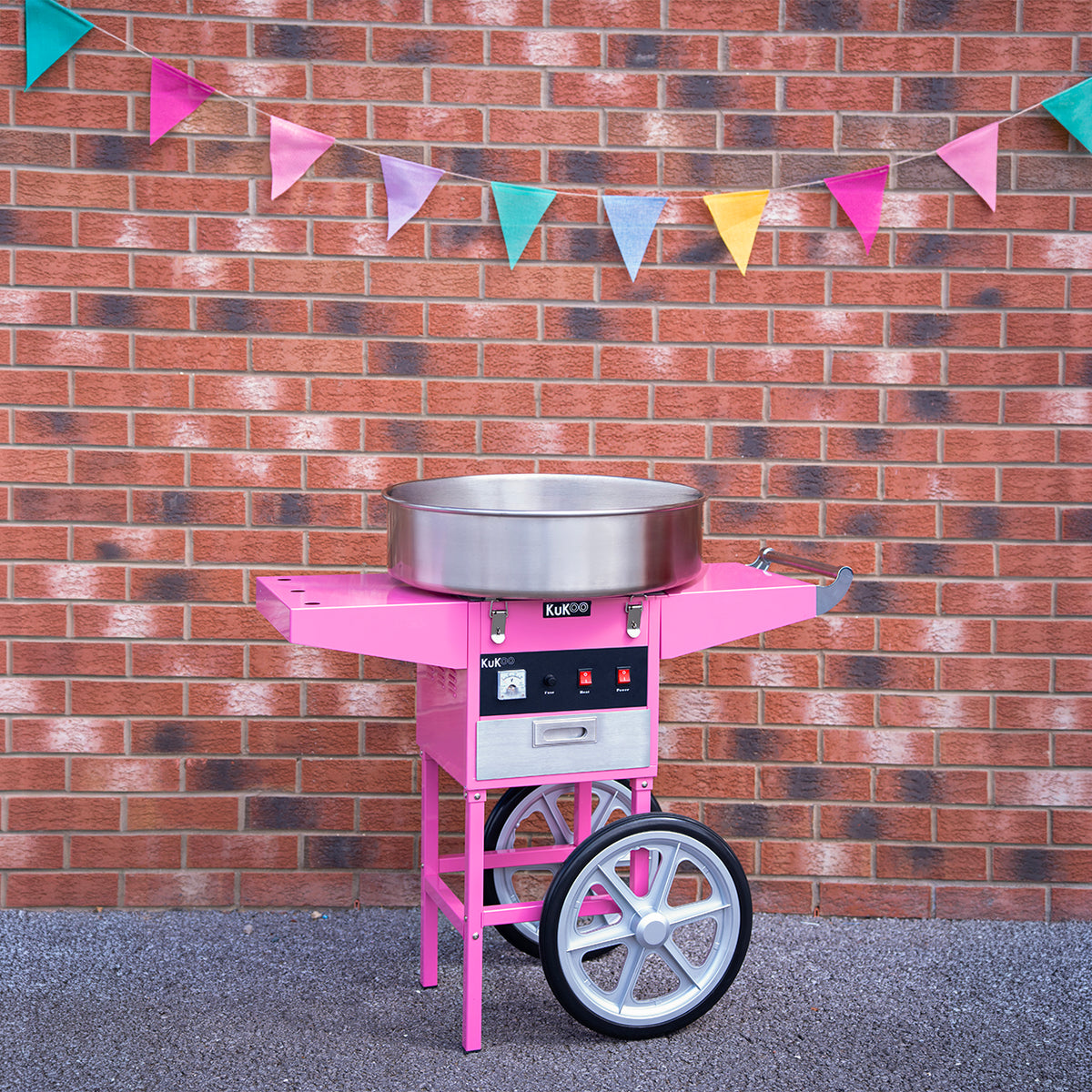 Bright pink cotton candy machine with stainless steel bowl, set against a brick wall and colorful triangular bunting