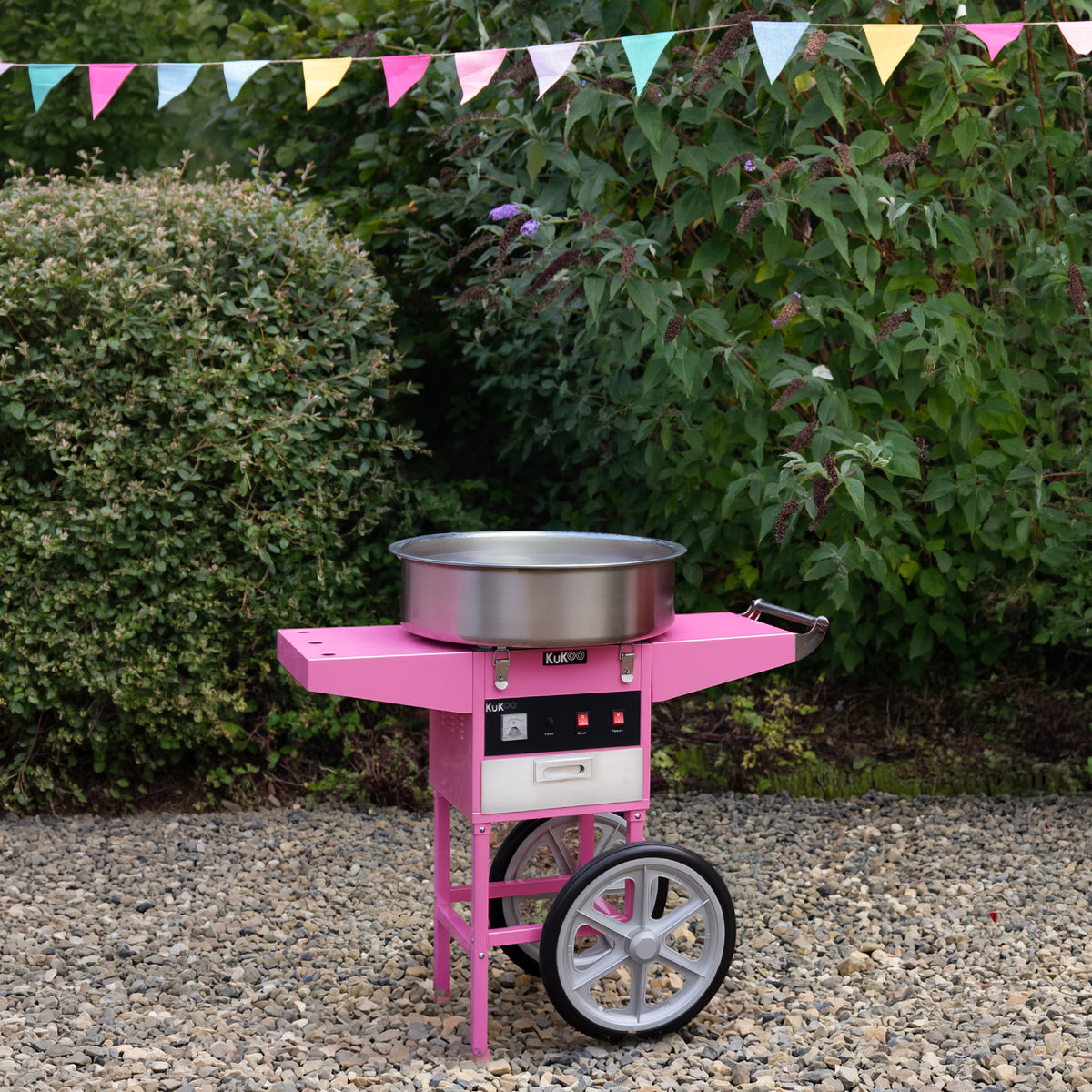 Bright pink cotton candy machine on a wheeled cart, set on gravel with greenery and colorful bunting in the background