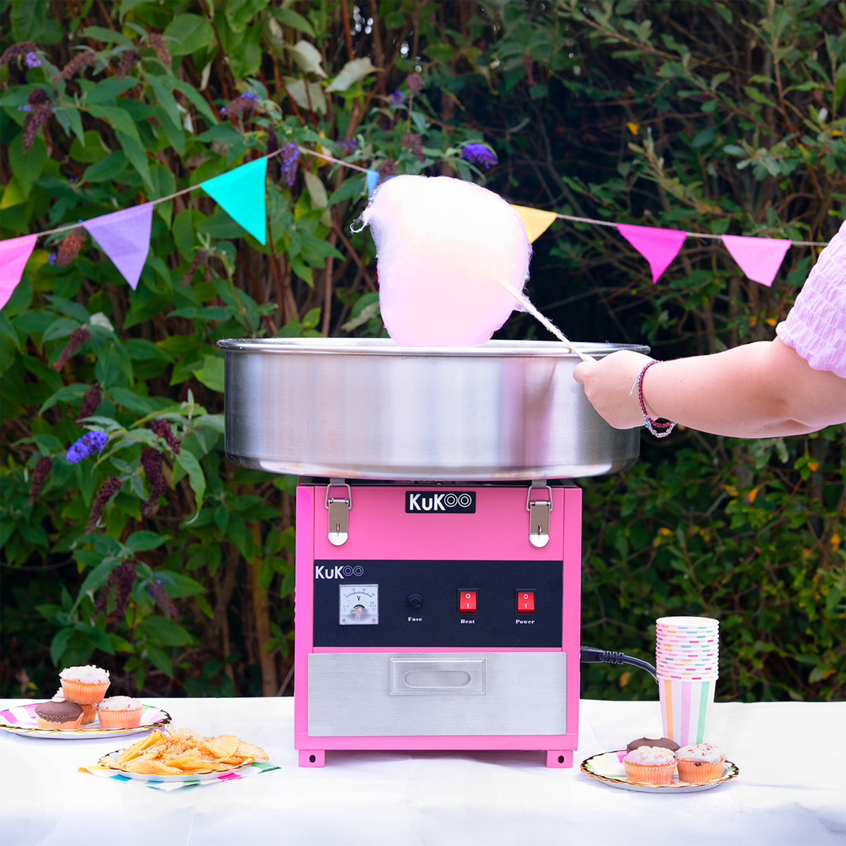 Cotton candy machine with a shiny silver bowl, pink base, and colorful party decorations, surrounded by cupcakes and cups