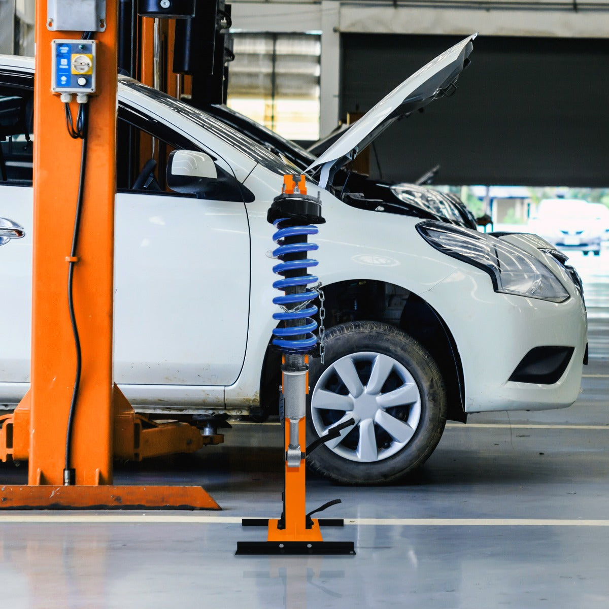Hydraulic coil spring strut in blue and orange, positioned beside a white car in a garage with an open hood, showcasing its sturdy design