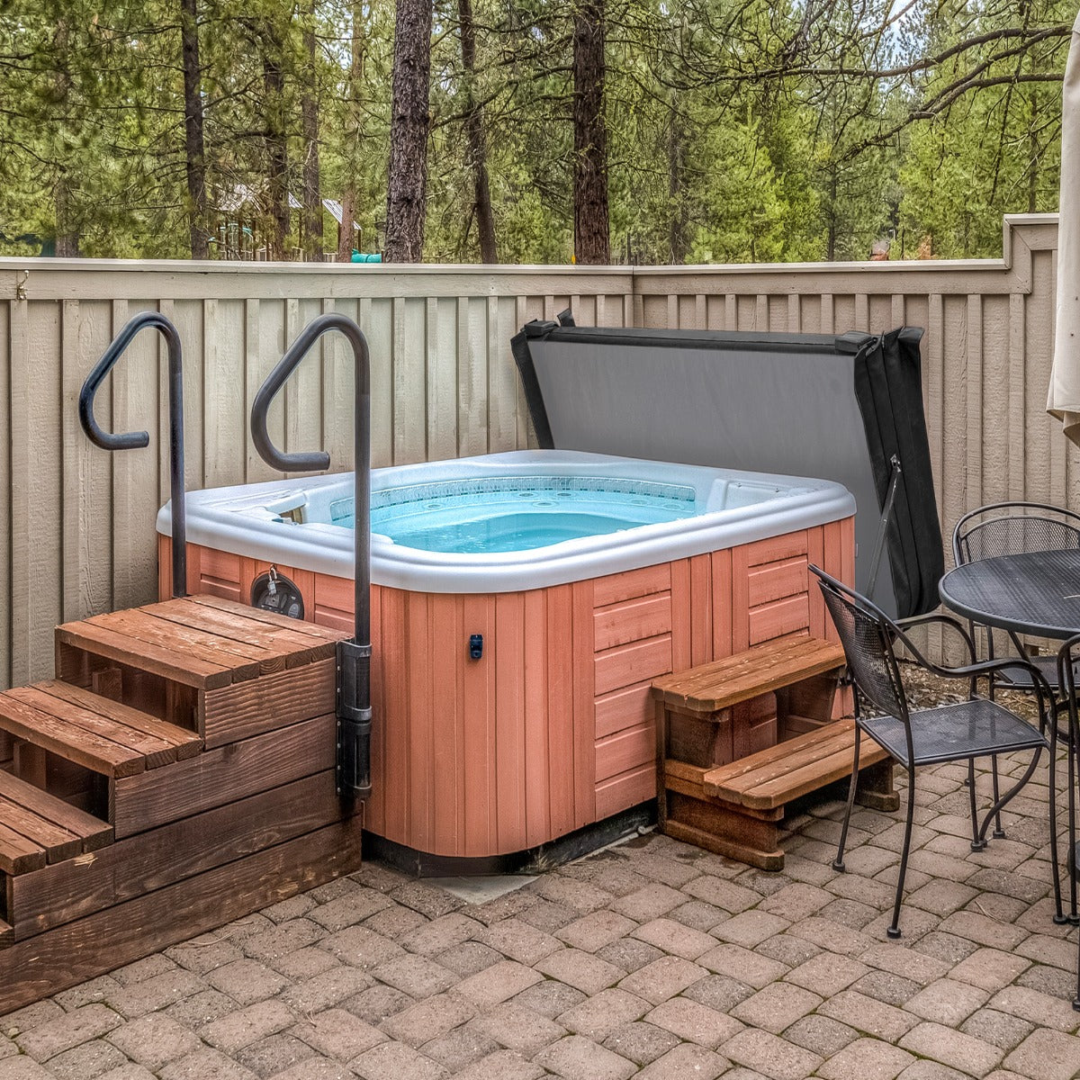 Black 2m hot tub spa cover resting on a wooden deck, surrounded by trees and a stone patio with outdoor seating