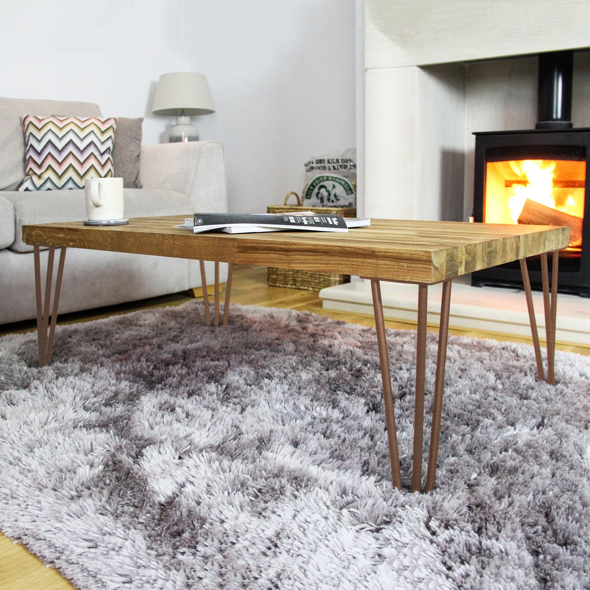Antique copper hairpin legs with a sleek 2-prong design support a wooden coffee table, set on a plush gray rug near a cozy fireplace