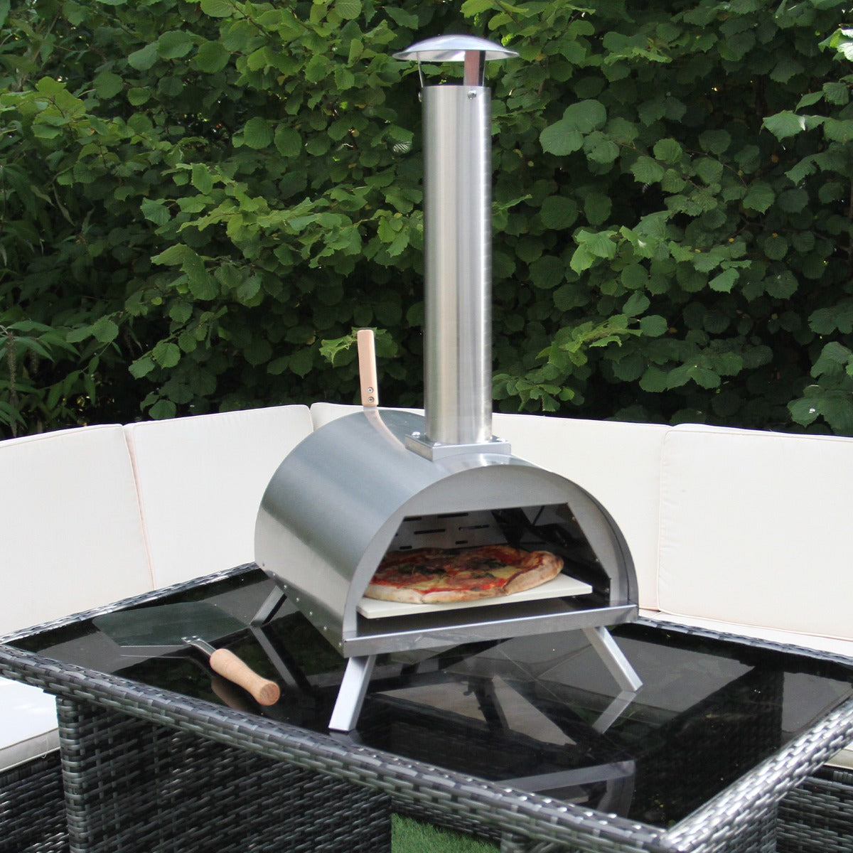 Stainless steel outdoor pizza oven with a chimney, cooking a pizza on a glass-top table surrounded by greenery