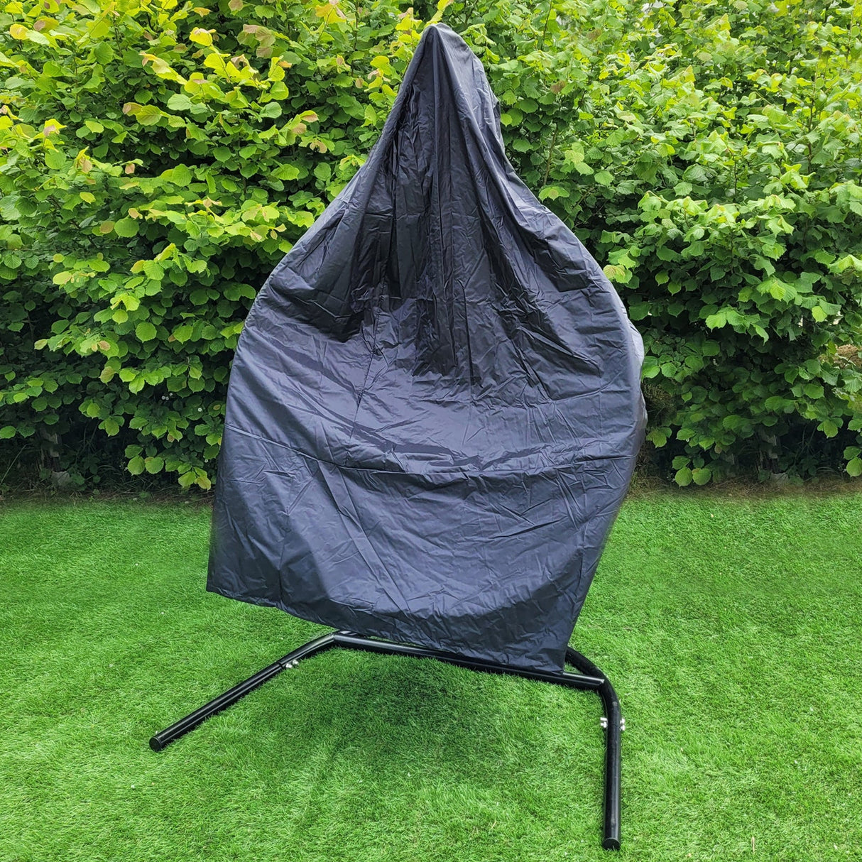 Two black hanging rattan egg chairs covered with protective tarps, set against a lush green hedge on a grassy surface