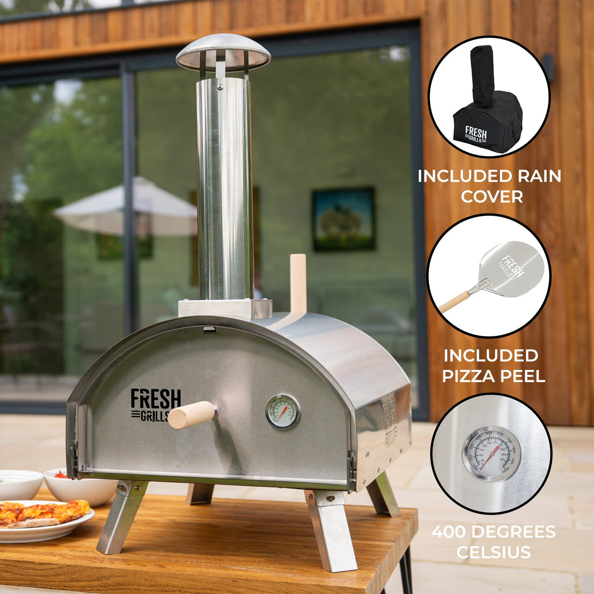 Premium stainless steel outdoor pizza oven with a chimney, thermometer, and included pizza peel and rain cover, set on a wooden patio