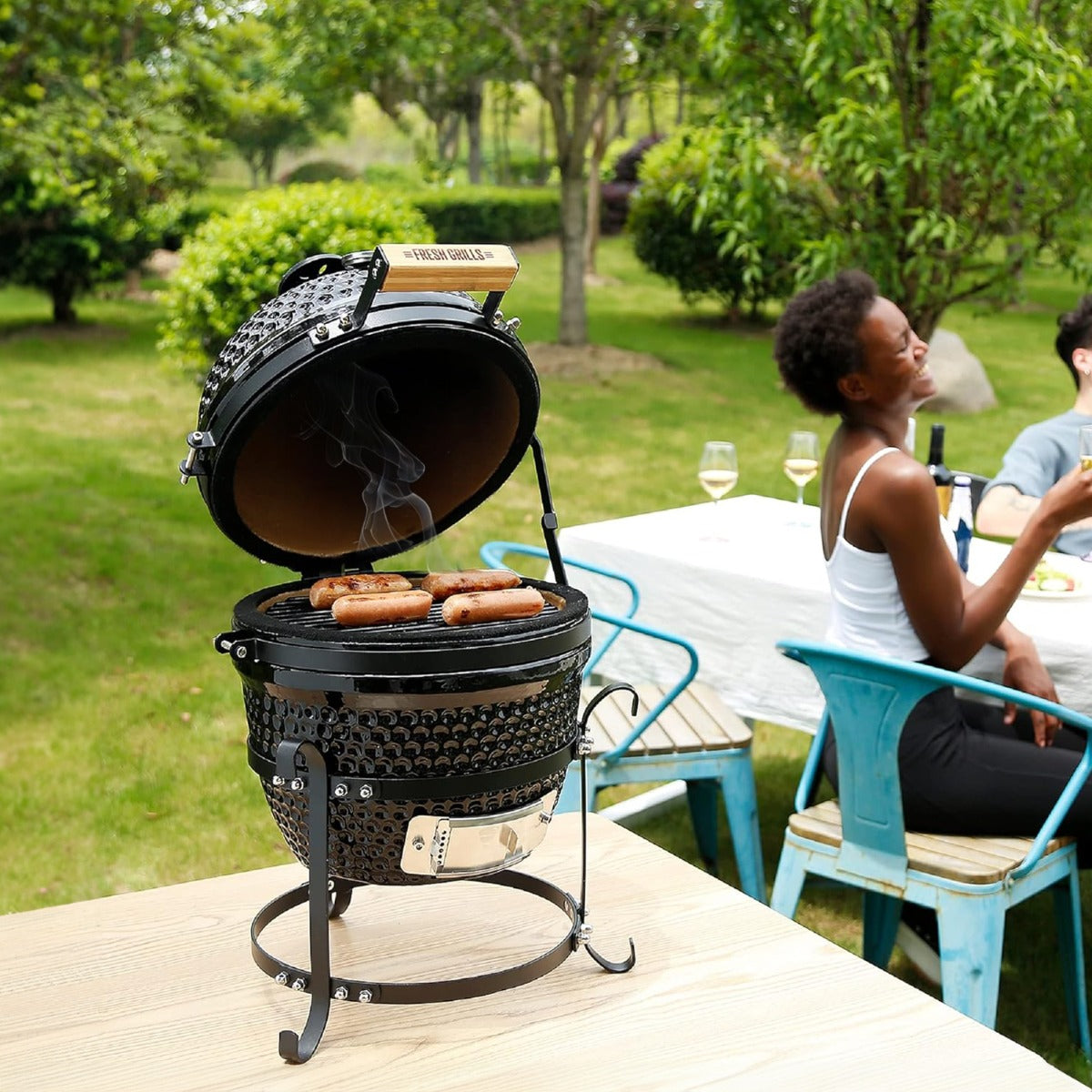 Black 13" Kamado BBQ oven with smoke rising, grilling sausages outdoors, surrounded by green trees and a dining setup