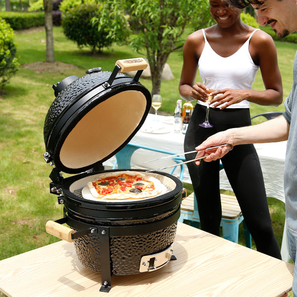 Black 16” Kamado BBQ oven with a textured finish, open lid revealing a pizza inside, set in a sunny outdoor garden scene