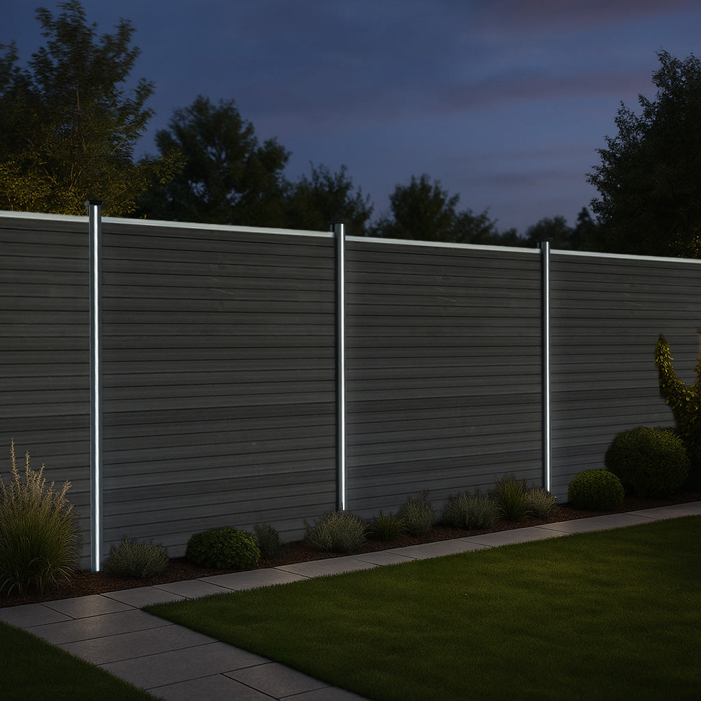 Grey composite fence panels with sleek LED posts, illuminated at night, surrounded by lush greenery and a stone pathway