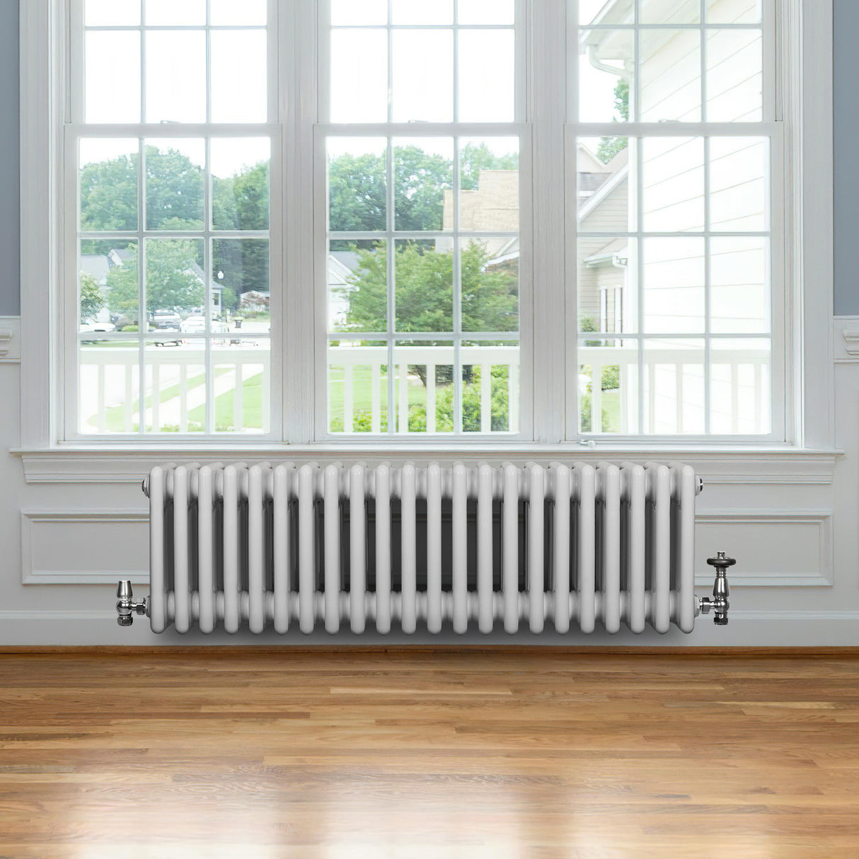 Traditional white 4 column radiator with valves, featuring a sleek design, installed beneath a large window in a bright room with wooden flooring