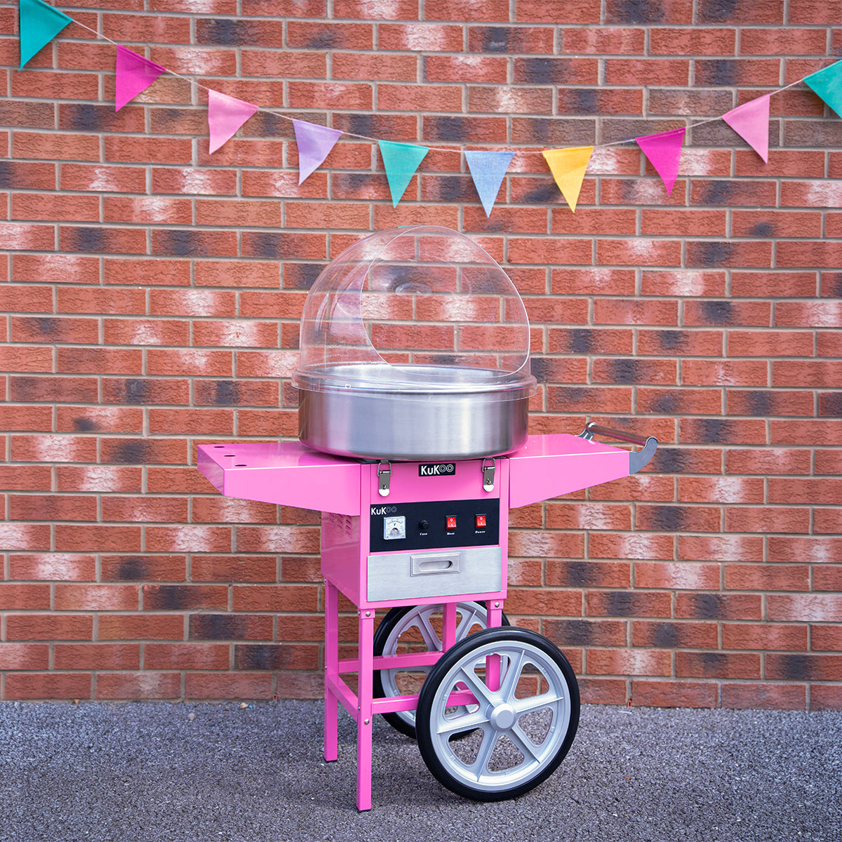Bright pink cotton candy machine with a shiny stainless steel bowl, set against a brick wall and colorful bunting flags