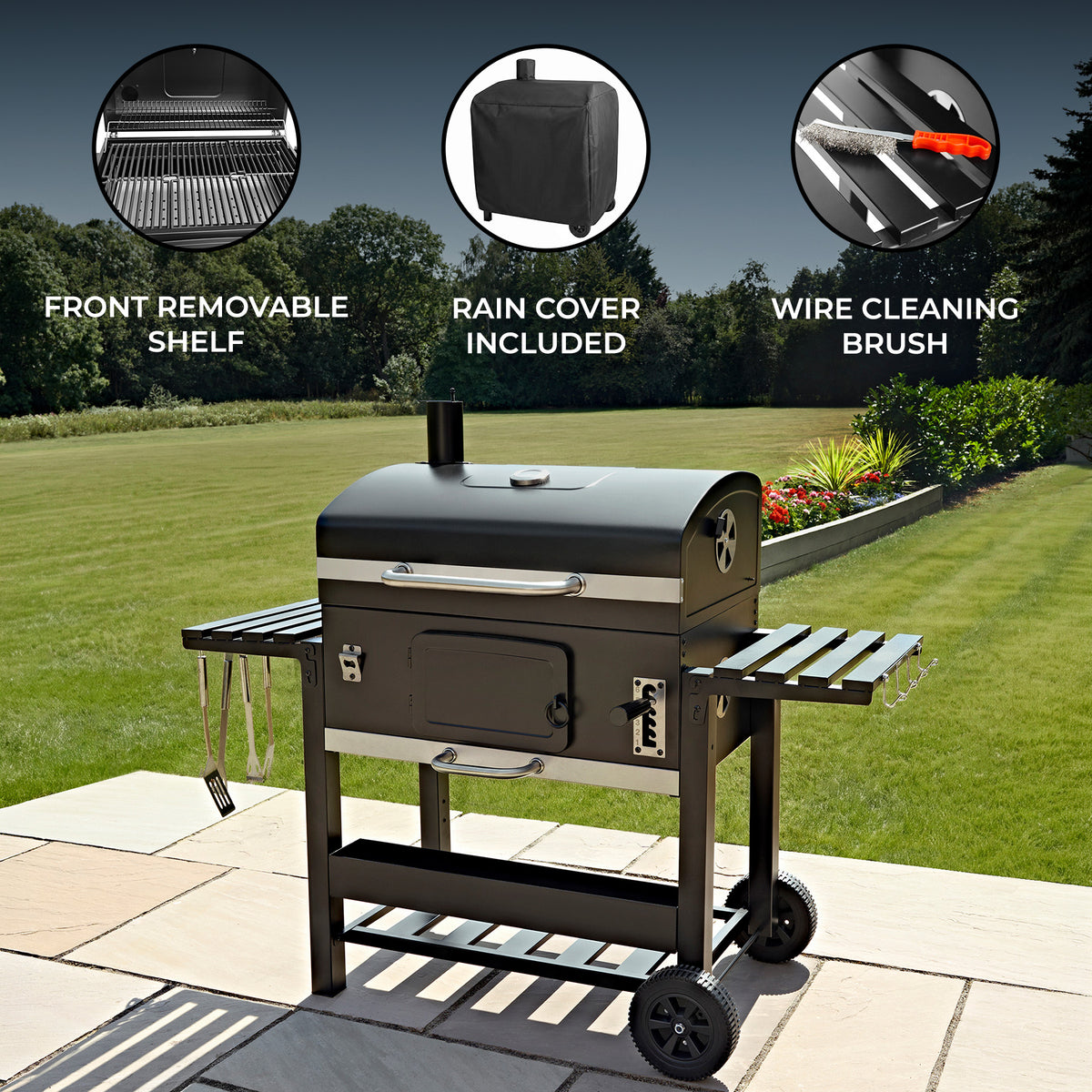 Black outdoor grill with a front removable shelf, rain cover, and wire cleaning brush, set on a patio with green grass in the background