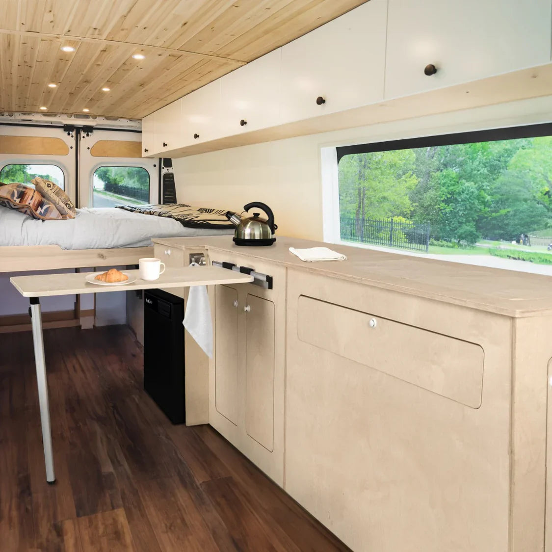 Cozy camper van interior featuring a wooden ceiling, sleek kitchen area, foldable table, and a bed with soft bedding, surrounded by greenery outside