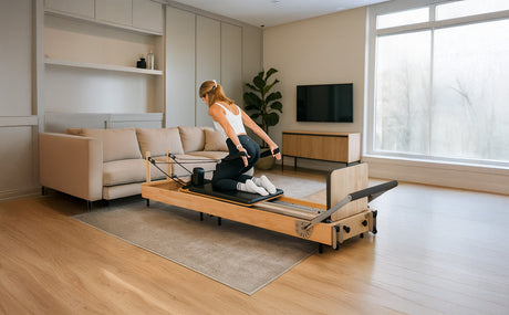 A woman performs Pilates on a wooden reformer in a modern, bright living room with a sofa and large window