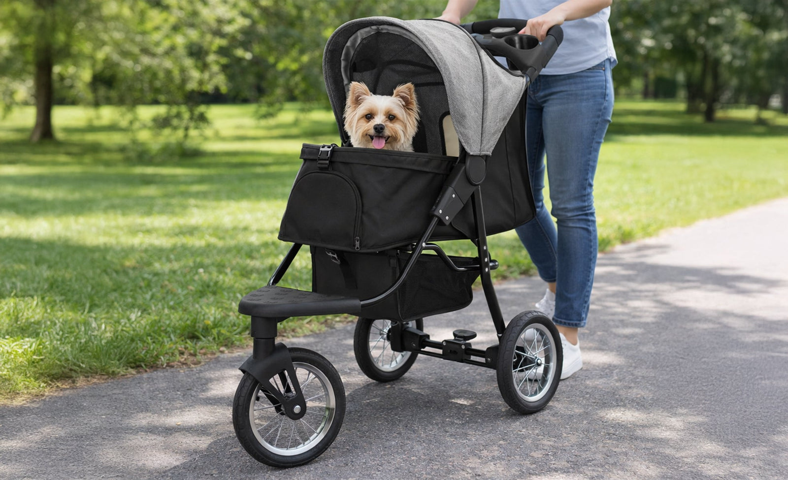 Stylish black pet jogger with a gray canopy, featuring a happy small dog inside, rolling on a path in a green park setting