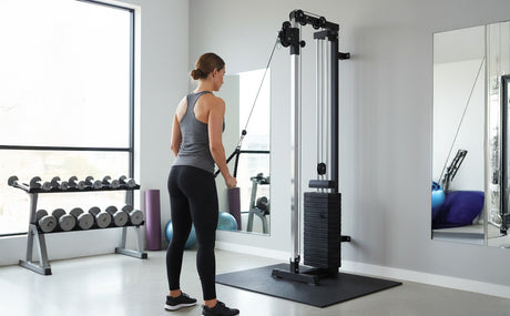 A woman in workout attire uses a wall-mounted cable machine in a modern gym, featuring weights and exercise equipment in the background