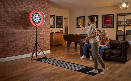 A woman throws a dart at a red freestanding dartboard in a cozy game room with a brick wall and a leather sofa