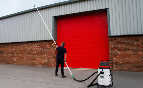 A person using a long pole vacuum to clean gutters, with a red industrial door and brick wall in the background