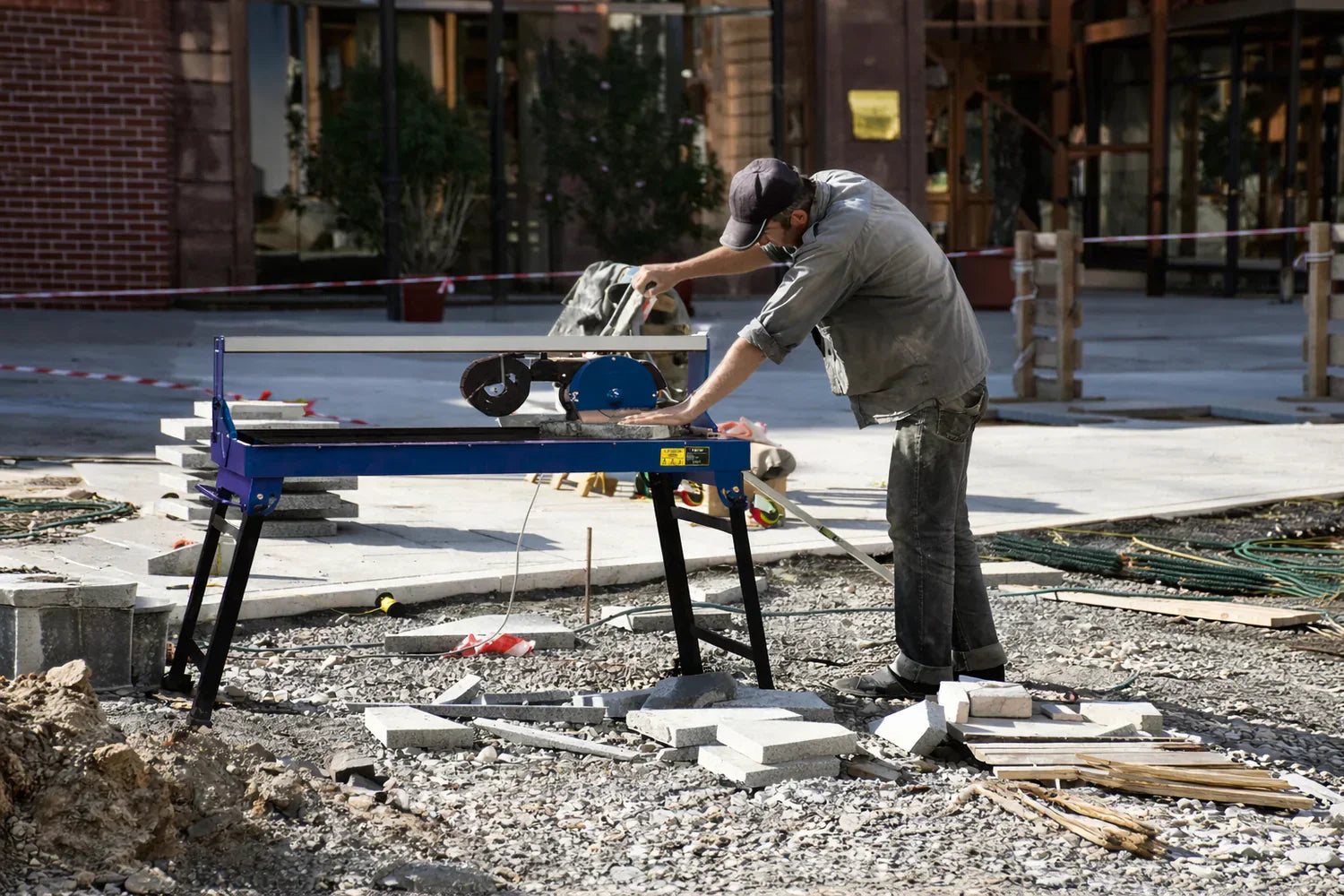 A worker using a wet tile cutter on a construction site, surrounded by gravel, tools, and building materials in a sunny environment
