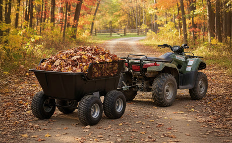A sturdy black trailer filled with colorful autumn leaves, attached to a green ATV, on a dirt path surrounded by vibrant fall foliage