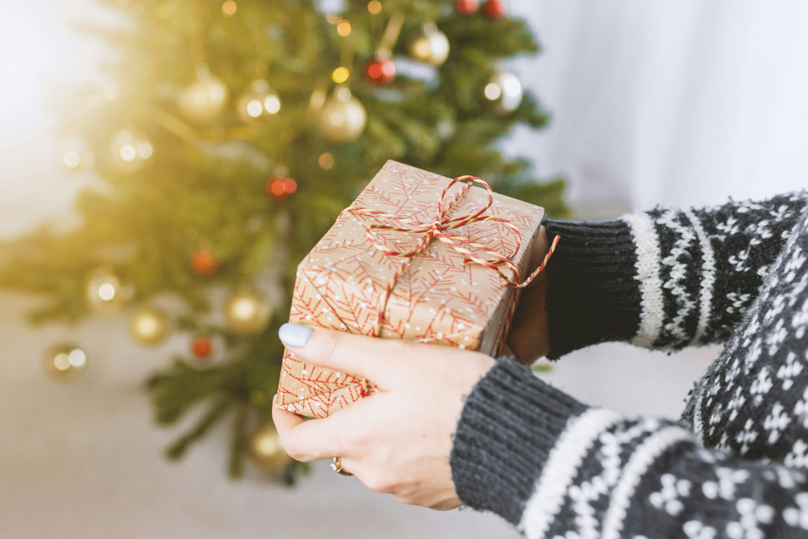A person in a cozy sweater holds a beautifully wrapped gift with a red bow, in front of a decorated Christmas tree