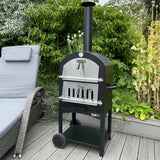 Black KuKoo outdoor pizza oven with stainless steel door, positioned on a wooden deck surrounded by greenery and a lounge chair