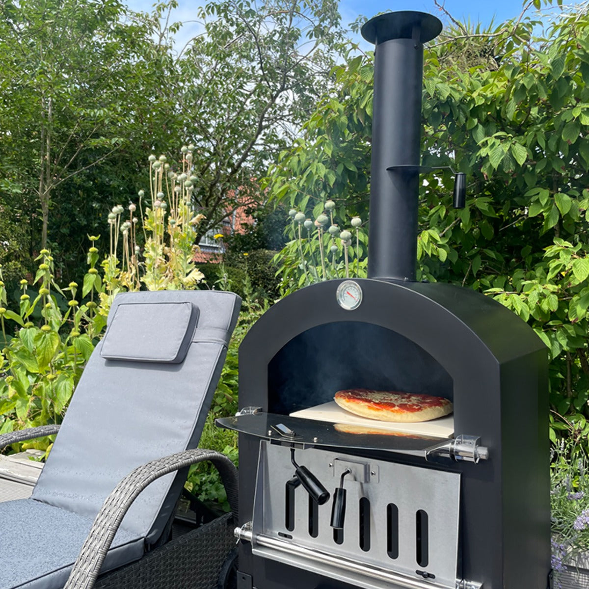 Black KuKoo outdoor pizza oven with a pizza baking inside, surrounded by greenery and a gray lounge chair in a sunny garden setting