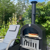 Black KuKoo outdoor pizza oven with a pizza baking inside, surrounded by greenery and a gray lounge chair in a sunny garden setting