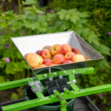 Stainless steel fruit crusher with a green frame, filled with red apples, ready for juicing or cider making in a garden setting