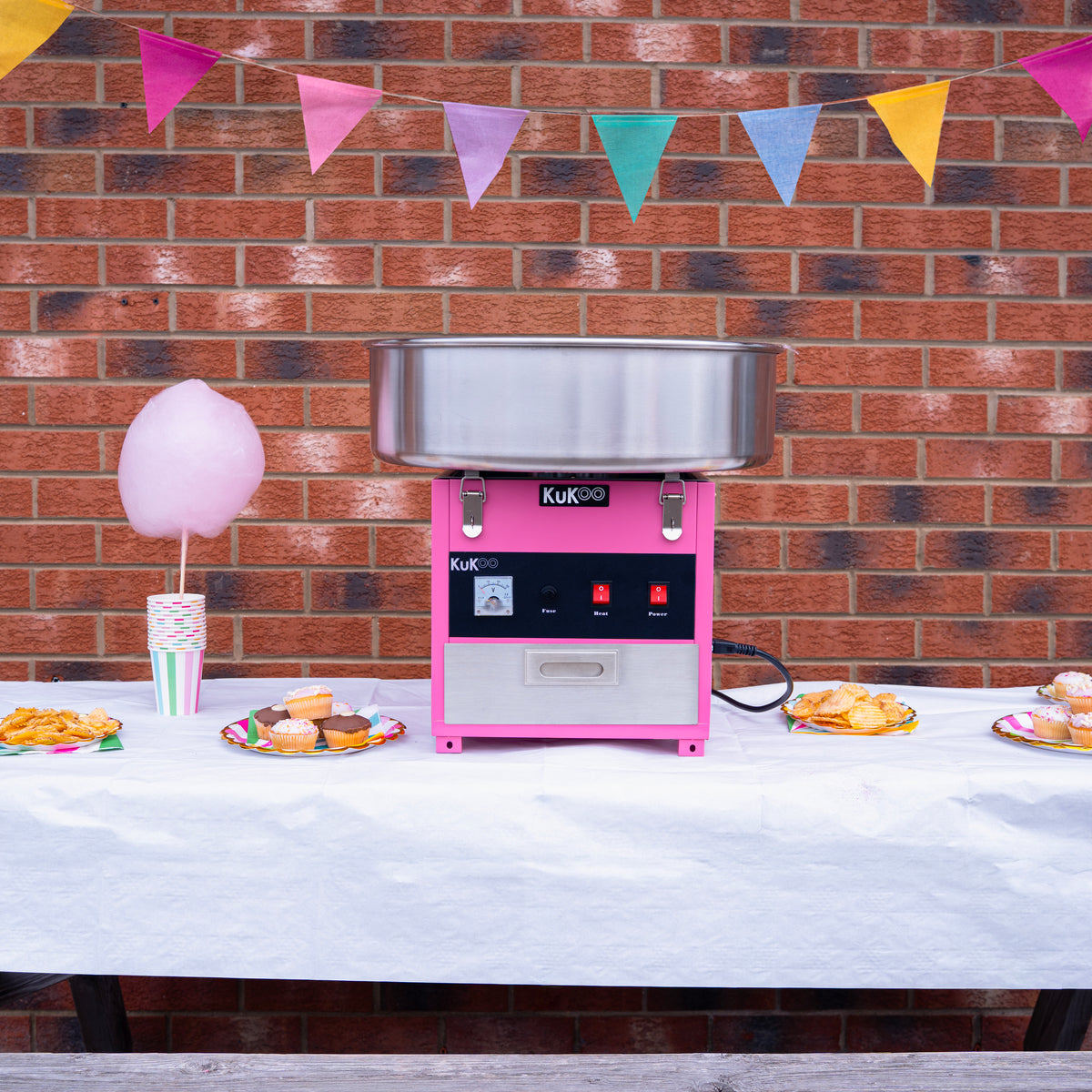 Cotton candy machine in pink with stainless steel bowl, set against a brick wall, surrounded by colorful party decorations and treats