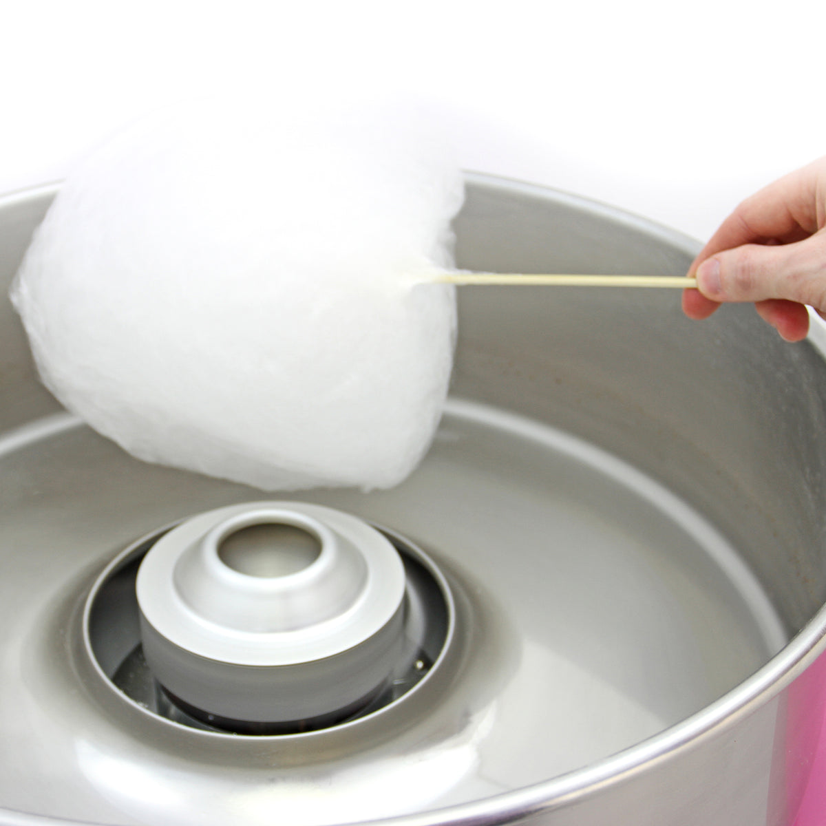 Fluffy white cotton candy being held on a wooden stick above a shiny silver cotton candy machine with a pink base