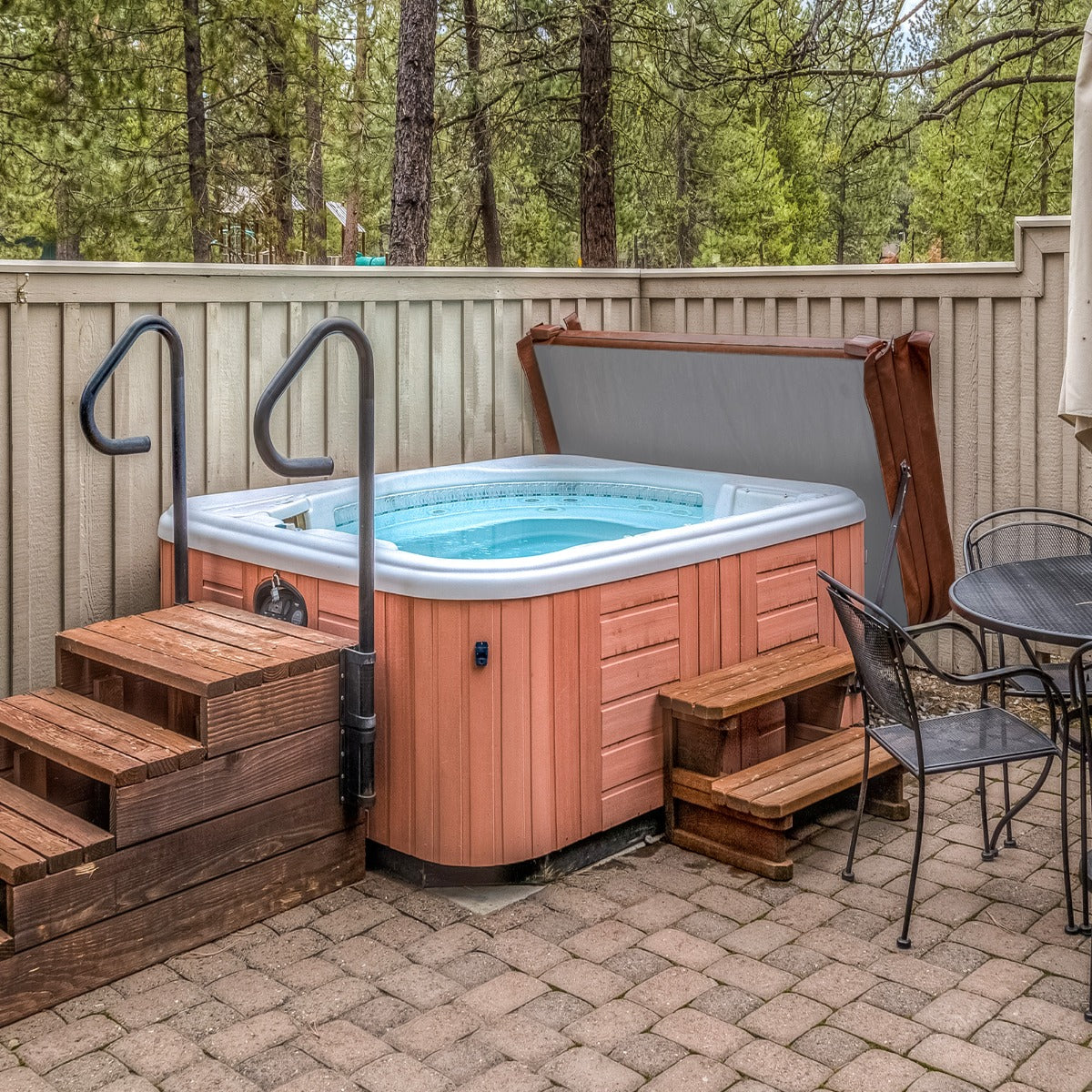 Brown 2m hot tub spa cover resting on a wooden deck, surrounded by trees, with steps leading up to the spa and a patio table nearby