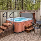 Brown 2m hot tub spa cover resting on a wooden deck, surrounded by trees, with steps leading up to the spa and a patio table nearby
