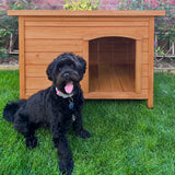 Small wooden dog kennel with a warm brown finish, featuring an arched entrance, placed on green grass with a black dog sitting nearby