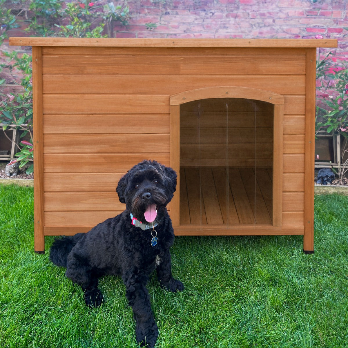 Large wooden dog kennel with a slanted roof, featuring a spacious entrance, set on green grass with a playful black dog in front