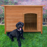 Large wooden dog kennel with a slanted roof, featuring a spacious entrance, set on green grass with a playful black dog in front
