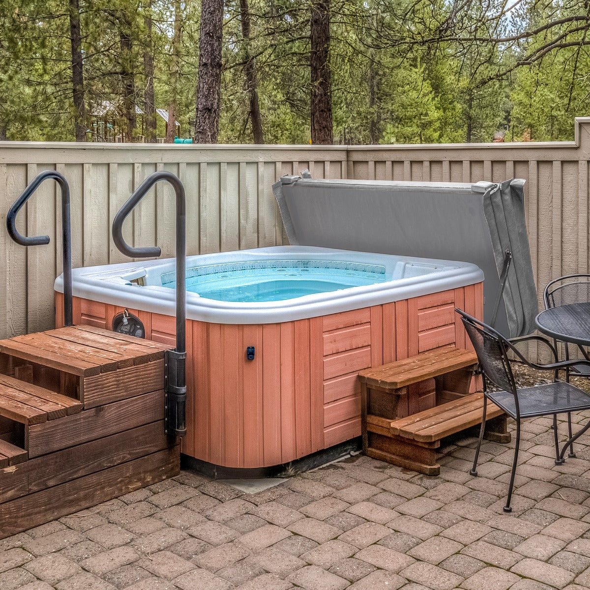 Grey 2.1m hot tub spa cover resting on a wooden hot tub, surrounded by trees and a stone patio with wooden steps and black chairs