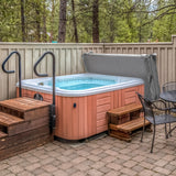 Grey 2.1m hot tub spa cover resting on a wooden hot tub, surrounded by trees and a stone patio with wooden steps and black chairs