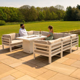 Modular white garden furniture set with soft beige cushions and a central fire pit, surrounded by lush green landscaping and blue sky