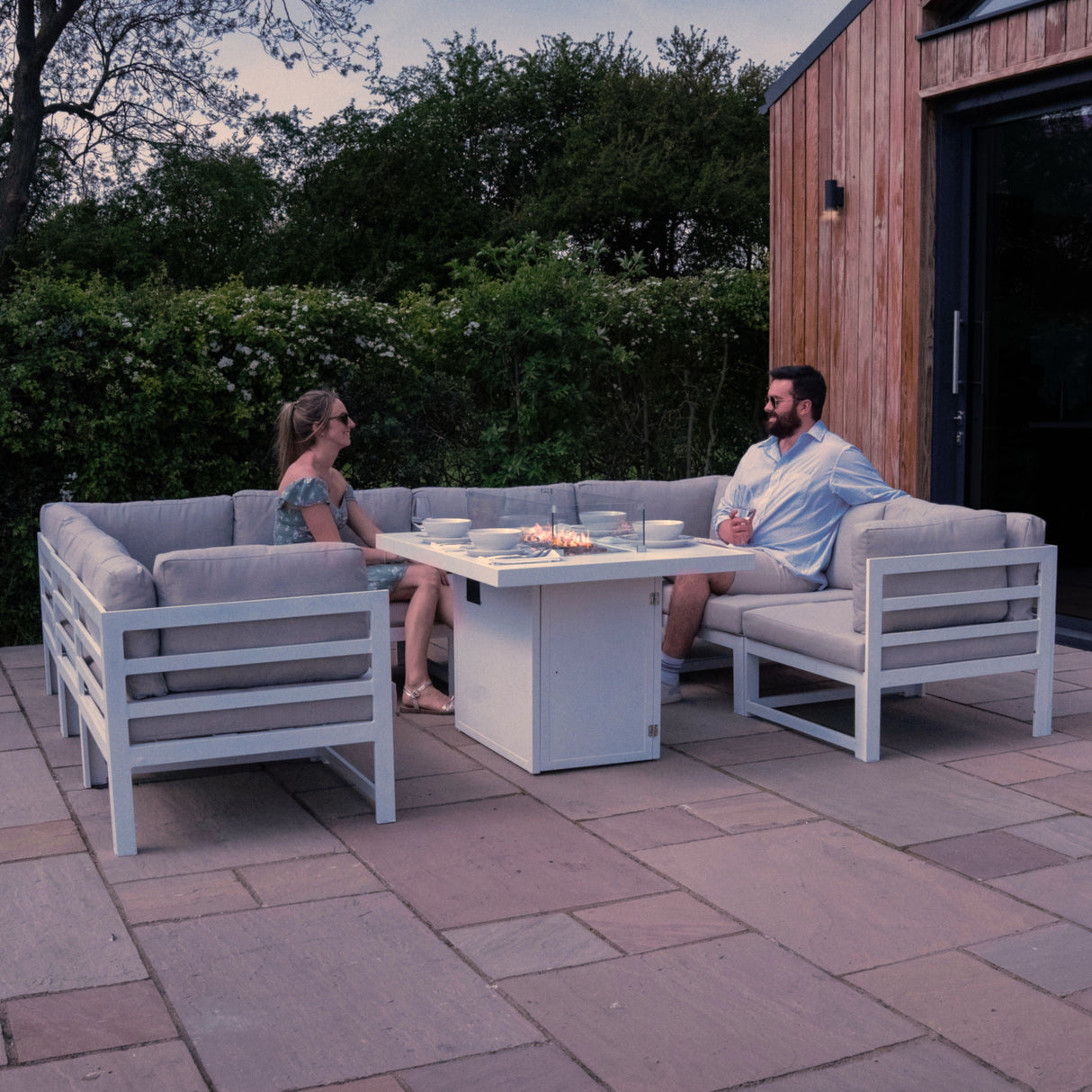 Modular white garden furniture set with plush cushions, featuring a central fire pit, surrounded by greenery and stone patio