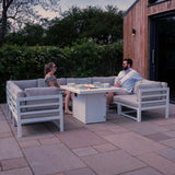 Modular white garden furniture set with plush cushions, featuring a central fire pit, surrounded by greenery and stone patio