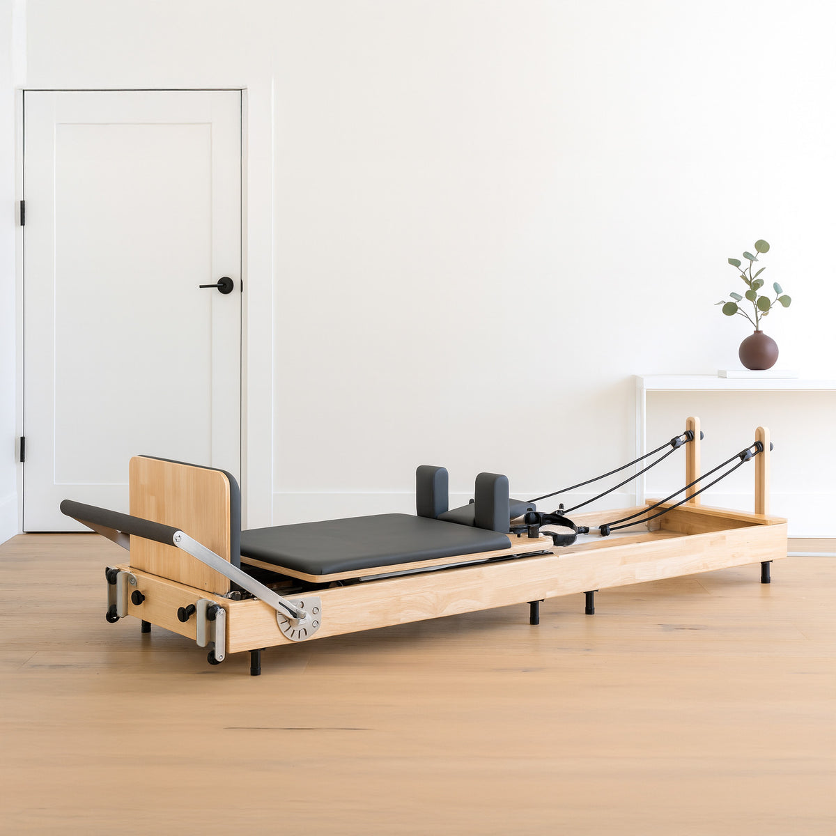 Wooden Pilates reformer with a black padded platform, adjustable springs, and a minimalist design, set in a bright, airy room