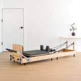 Wooden Pilates reformer with a black padded platform, adjustable springs, and a minimalist design, set in a bright, airy room