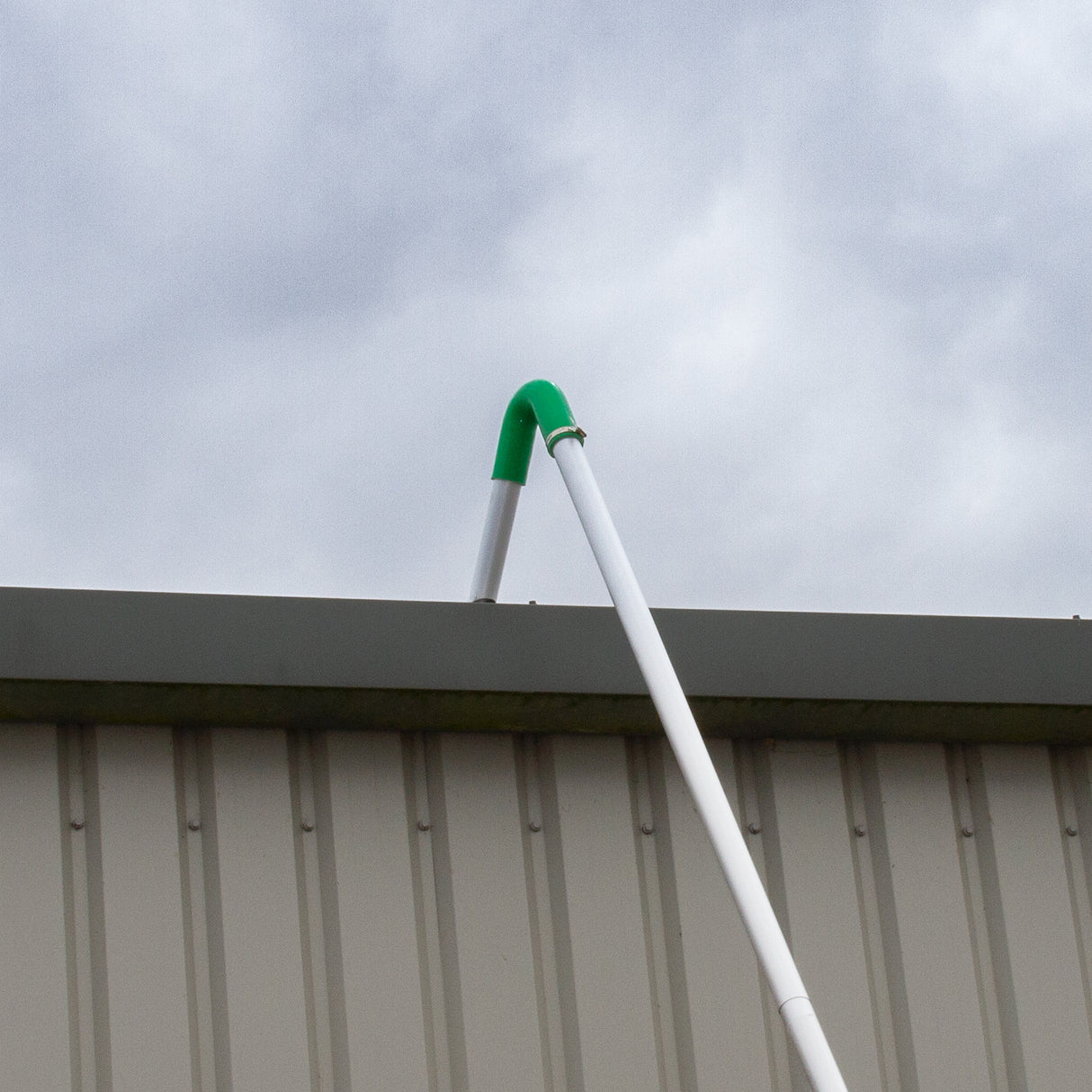 White gutter vacuum pole with a green curved attachment, positioned against a gray metal building under a cloudy sky