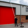A person using a 80L wet and dry vacuum with a long pole to clean gutters, featuring a red building backdrop and gray pavement