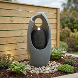 Sleek gray electric water feature with cascading falls, surrounded by smooth pebbles and lush green plants, enhancing outdoor aesthetics