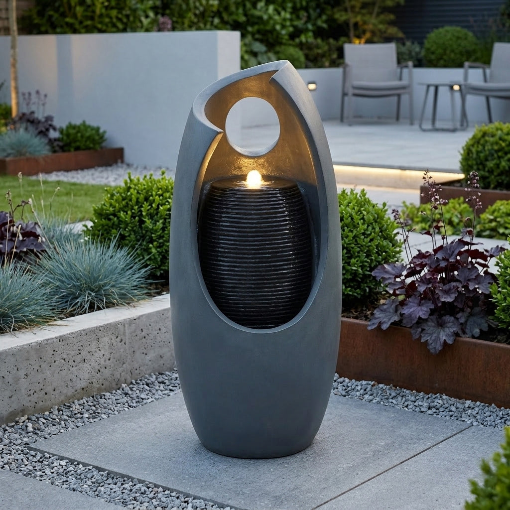 Sleek gray electric water feature with a circular opening, cascading water, and soft glow, surrounded by lush greenery and modern landscaping
