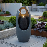 Sleek gray electric water feature with a circular opening, cascading water, and soft glow, surrounded by lush greenery and modern landscaping