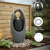 Sleek gray electric water feature with ambient LED lighting, cascading water, and a modern design, surrounded by decorative stones and plants