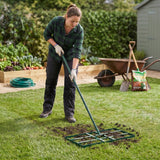A woman using a green 100cm lawn leveller on a grassy yard, surrounded by garden beds and gardening tools in the background